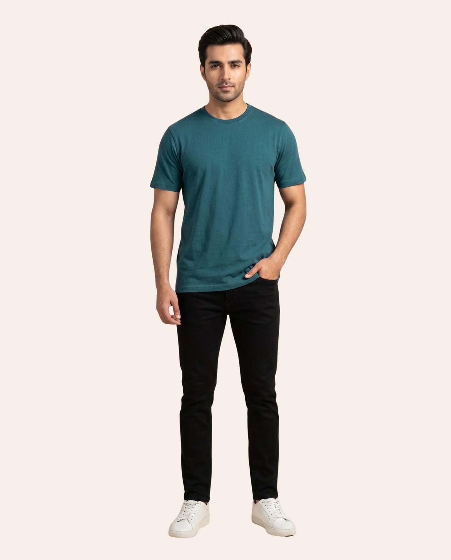 A man modeling a teal short-sleeve crewneck t-shirt, paired with black slim-fit jeans and white sneakers, standing against a solid light beige background.