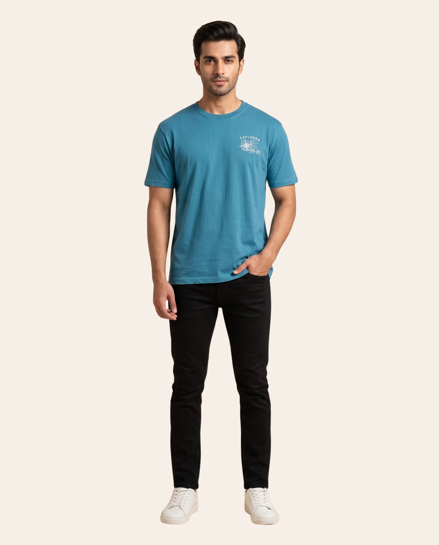 A man modeling a short-sleeve crew-neck t-shirt in a heathered teal blue color, paired with black jeans and white sneakers, against a solid light beige background.
