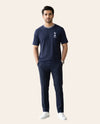 A man modeling a navy blue short-sleeve t-shirt and matching navy blue trousers. The t-shirt features a white embroidered crest on the left chest and a patterned collar.