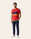 A man modeling a red short-sleeve t-shirt with navy and yellow horizontal stripes across the chest, paired with navy pants and white sneakers, against a solid pale beige background.