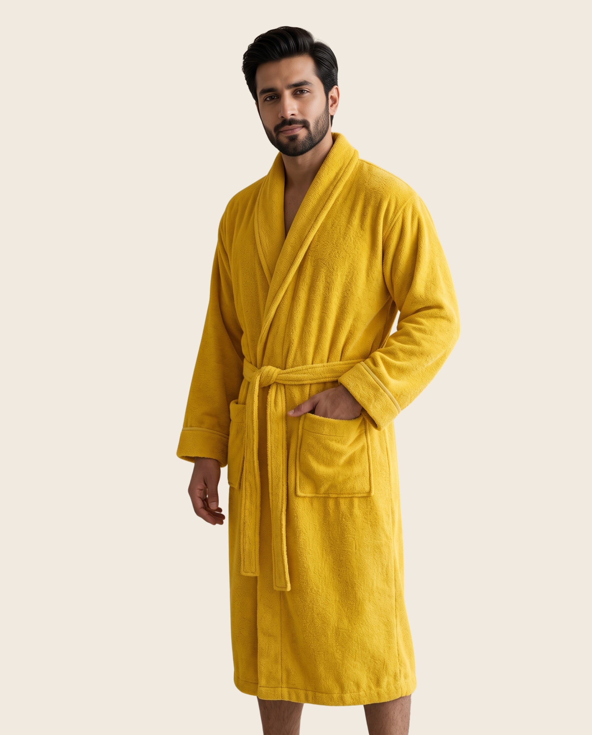 A man wearing a plush mustard yellow bathrobe with a tie belt and front patch pocket, standing against a solid light beige background.